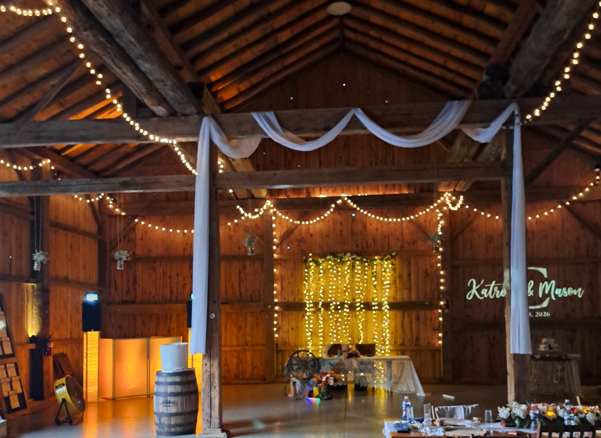 A barn reception before guests arrive. String lights run along the beams overhead, the walls are washed amber by uplighting, a curtain of fairy lights hangs behind the sweetheart table, and a monogram is projected on the far wall beside the lit DJ booth.