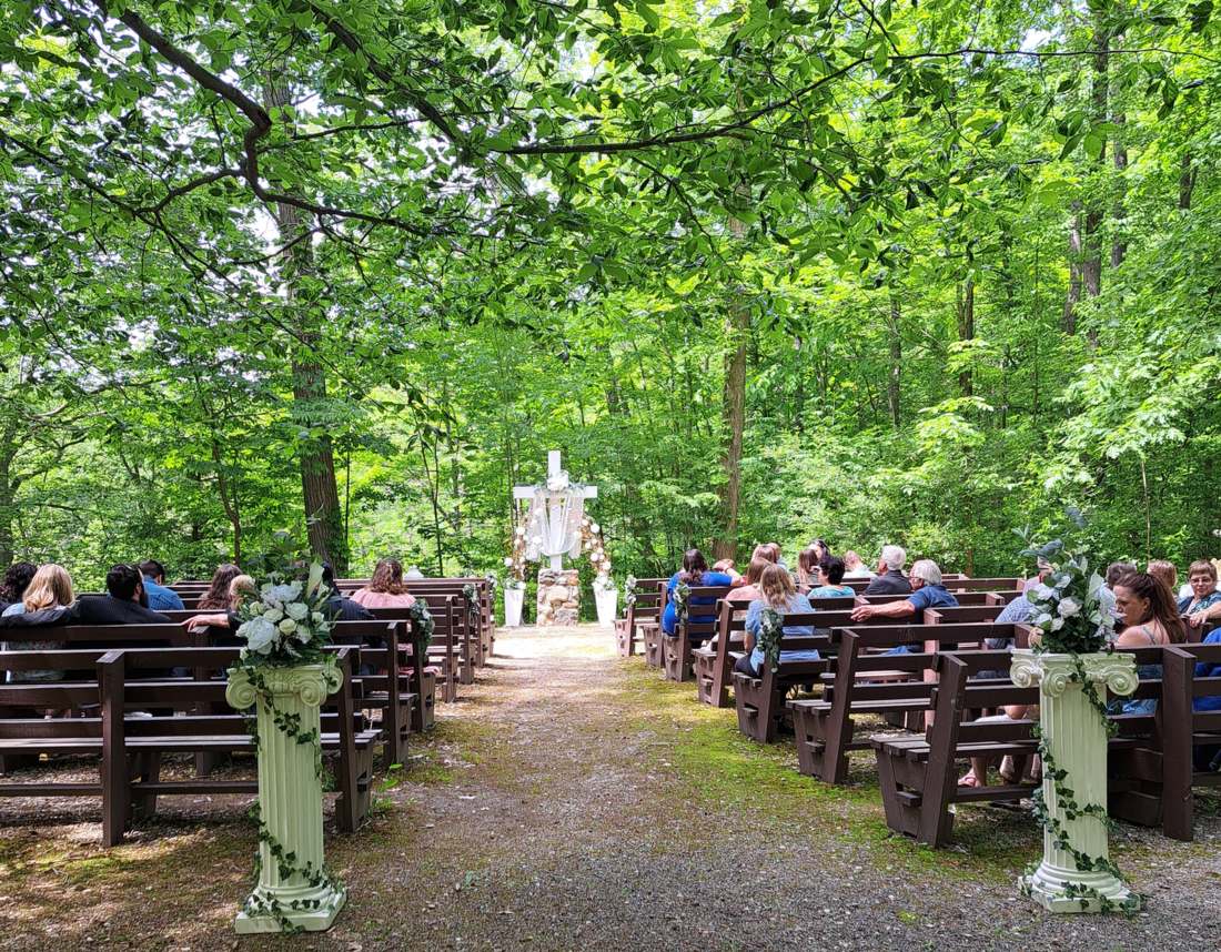 A wedding ceremony in a woodland chapel with no walls and no roof. Two ivy-wrapped columns topped with white roses mark the head of a gravel aisle, guests sit on dark wooden benches under a full green canopy, and a white cross draped in fabric stands at the far end. There is no building and no power line anywhere in sight.