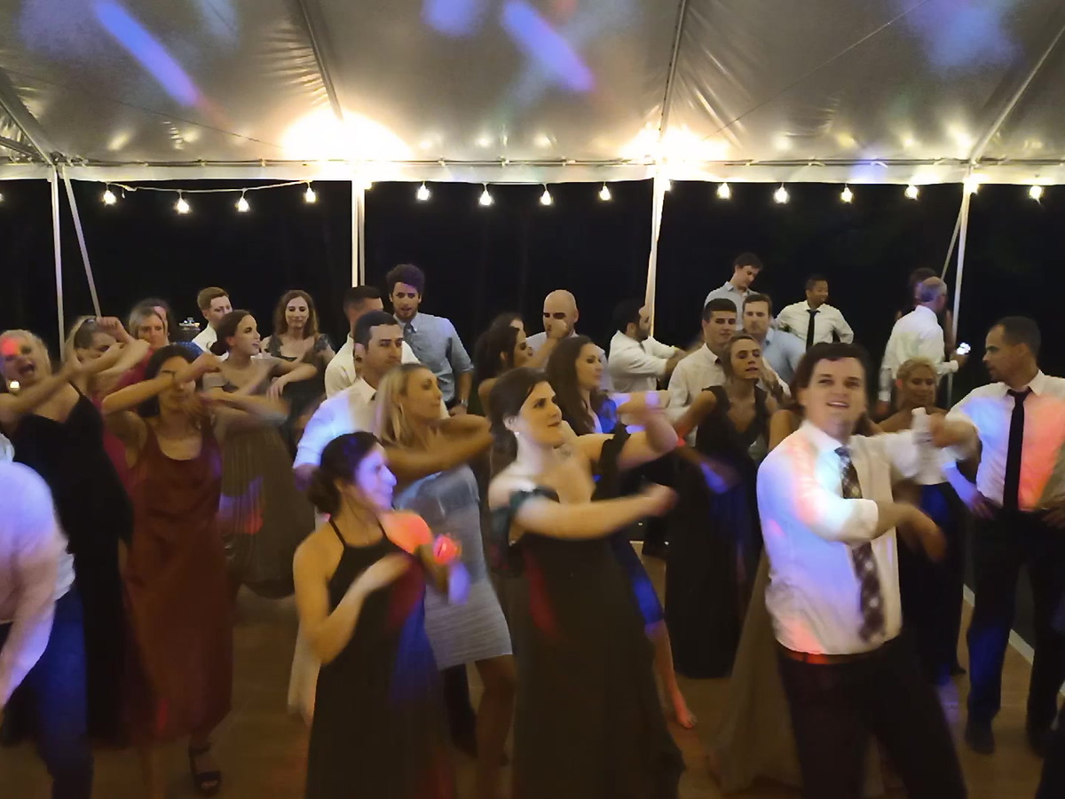 A packed dance floor under a tent at night. Around forty guests of all ages are caught mid-step in the same move, arms swung across their bodies, while string lights and colored stage light play across the white canopy overhead.