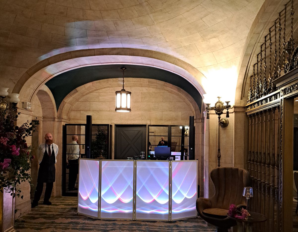 A DJ booth set up under a vaulted stone ceiling in a downtown Cleveland event space. The booth facade is backlit with overlapping arcs of pink, blue and white light, beside an ornate brass gate and a hanging lantern.