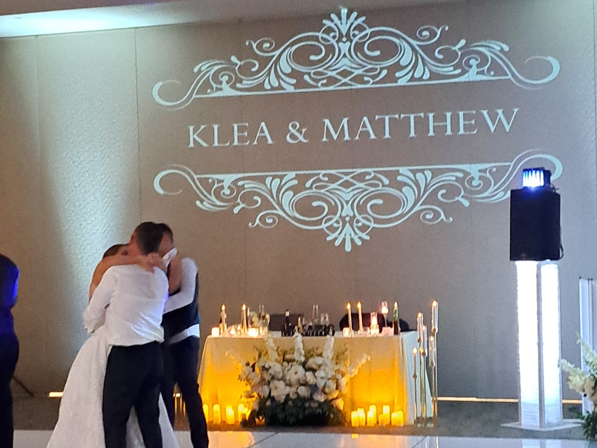 A couple during their first dance on a white floor, in front of a large custom monogram projected onto the ballroom wall above a candlelit sweetheart table.